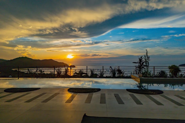 Sunset over ocean viewed from patio with pool and railing.