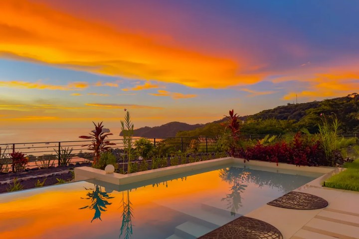 Vibrant sunset over mountains and pool with lush greenery.