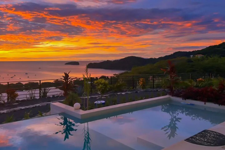 Ocean sunset view with pool and lush hills in the foreground.
