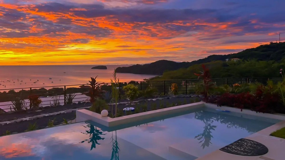Ocean sunset view with pool and lush hills in the foreground.