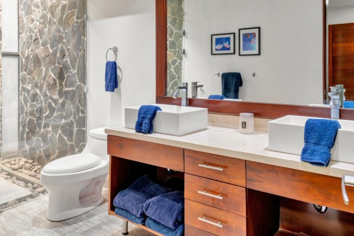 Modern bathroom with stone shower, double sinks, wooden vanity, and blue towels.