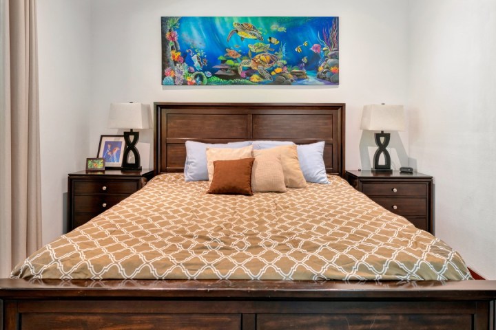 Bedroom with a wood bed, patterned duvet, pillows, side tables, lamps, and an ocean scene painting above the bed.