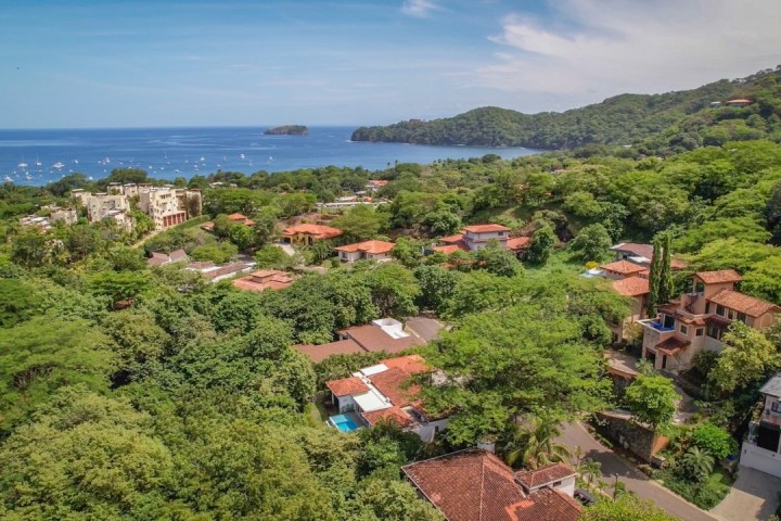 Aerial view of a coastal village with greenery, red-roofed houses, and ocean in the background.