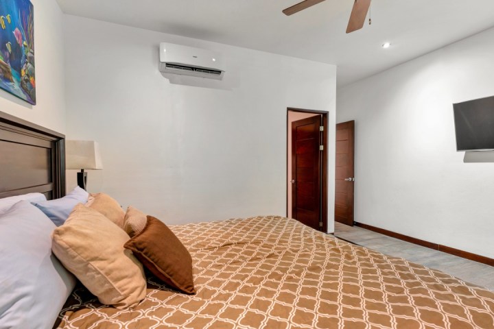 Bedroom with brown patterned bedspread, wall art, ceiling fan, air conditioner, and wall-mounted TV.