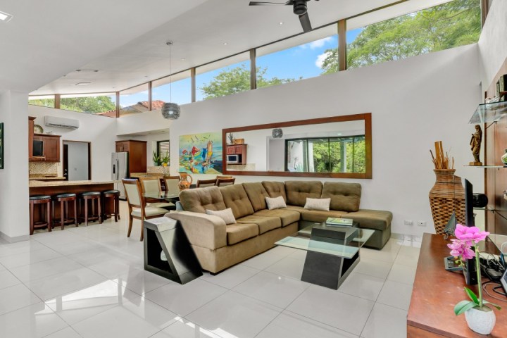 Modern living room with sectional sofa, glass coffee table, kitchen bar, and large windows overlooking trees.