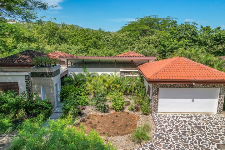 Aerial view of a house with red-tiled roofs, surrounded by lush greenery and trees.