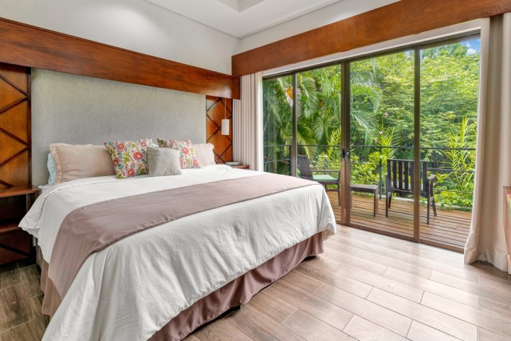 Bedroom with large bed, colorful pillows, and sliding glass doors to a balcony overlooking lush greenery.