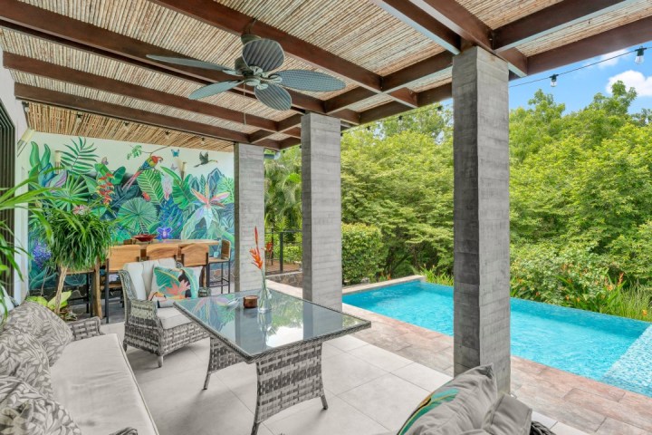 Outdoor patio with wicker furniture, mural, ceiling fan, pool, and lush greenery.