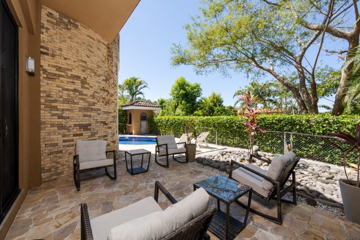 Patio with wicker chairs, table, and view of a pool and garden with trees under a clear blue sky.