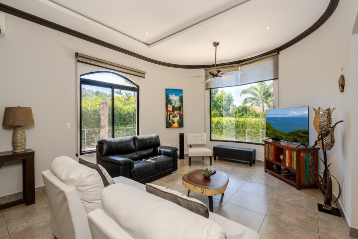 Modern living room with sofas, TV, ceiling fan, large windows, and a colorful painting.