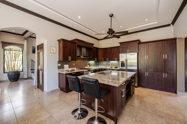 Modern kitchen with dark wood cabinets, granite island, two black chairs, and stainless steel appliances.