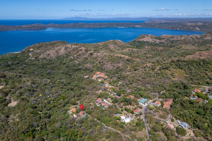 Aerial view of lush hills and coastline with scattered houses and a red location marker on a property.