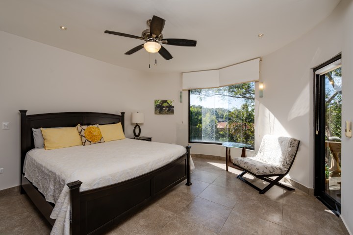 Modern bedroom with dark bed, yellow pillows, large window, chair, and ceiling fan.