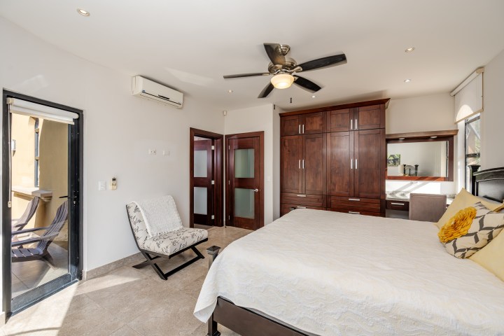 Modern bedroom with a bed, chair, wooden wardrobe, and ceiling fan near a glass door to a balcony.