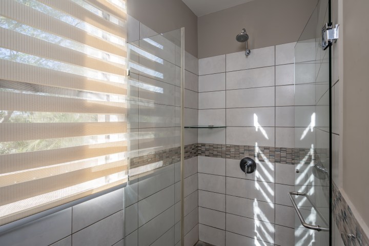 Glass shower with tiled walls and window blinds allowing sunlight in.