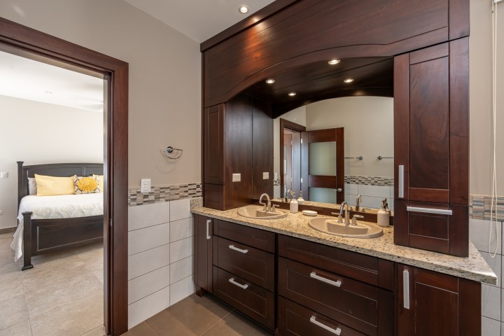 Bathroom with dual sinks, dark wood cabinets, and view of a bedroom with pillows on the bed.