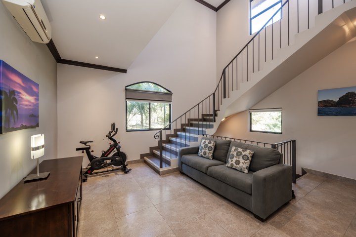Modern living room with a gray sofa, stationary bike, staircase, and large windows.