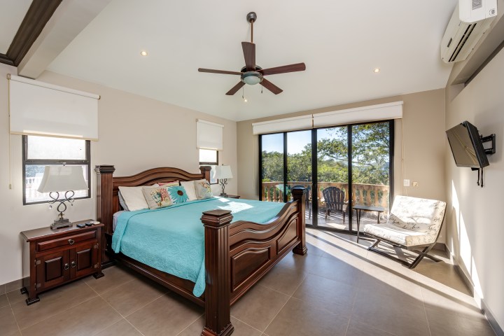 Spacious bedroom with a wooden bed, teal bedding, ceiling fan, and glass doors opening to a balcony with chairs.