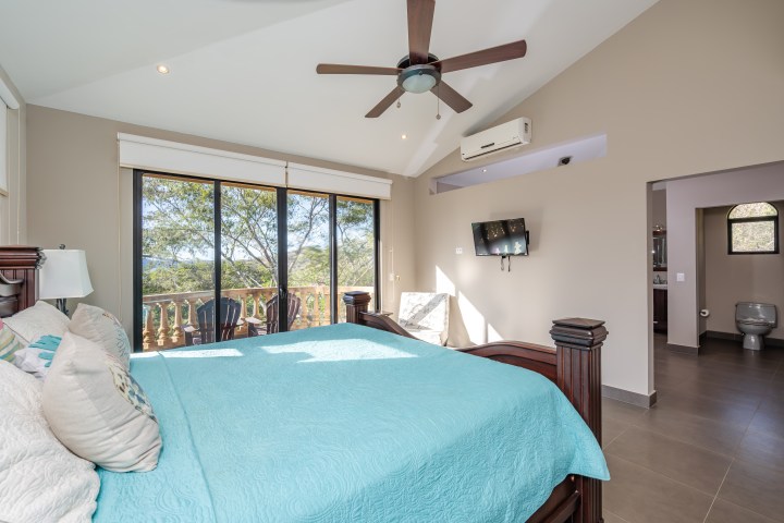 Bright bedroom with teal bedding, ceiling fan, TV, large window, and open bathroom view.