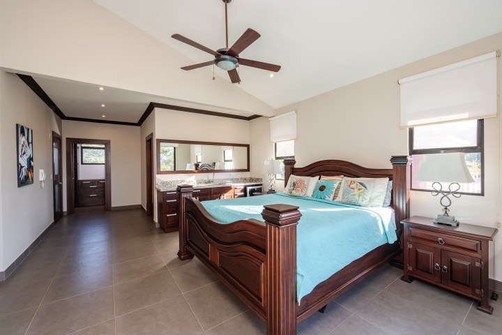 Spacious bedroom with wooden bed, blue bedding, ceiling fan, and large windows.