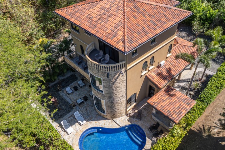 Aerial view of a villa with red-tiled roof, patio, and a blue kidney-shaped pool surrounded by greenery.