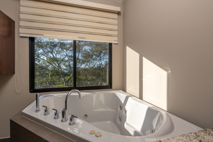 Bathroom with a large corner bathtub and a window with blinds.