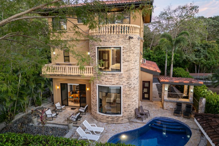Three-story house with pool, patio furniture, and trees surrounding.