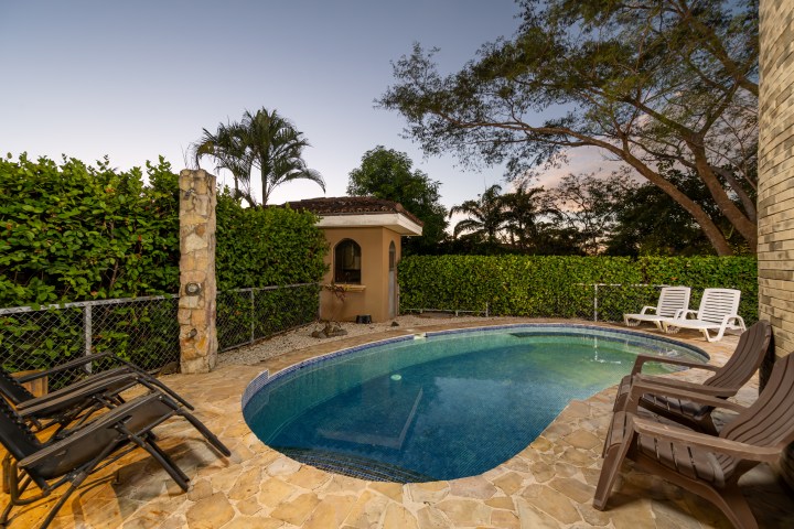 Small backyard pool with lounge chairs, surrounded by trees and a stone wall at dusk.