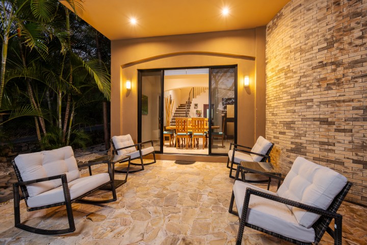 Outdoor patio with wicker chairs and cushioned seats, leading to indoor dining area through glass doors at dusk.