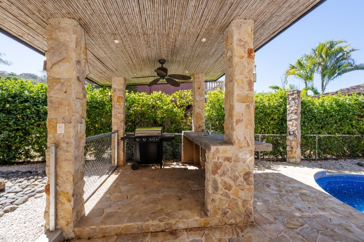 Outdoor kitchen with stone pillars, grill, and ceiling fan next to a pool and greenery.