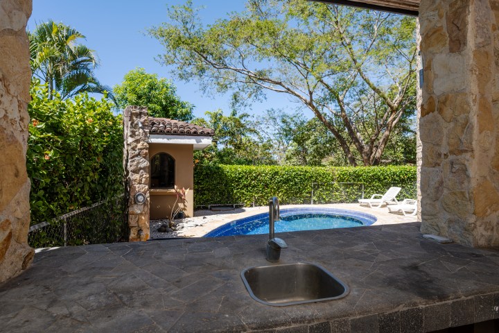 View from kitchen sink to pool, patio, and garden with trees under a clear blue sky.