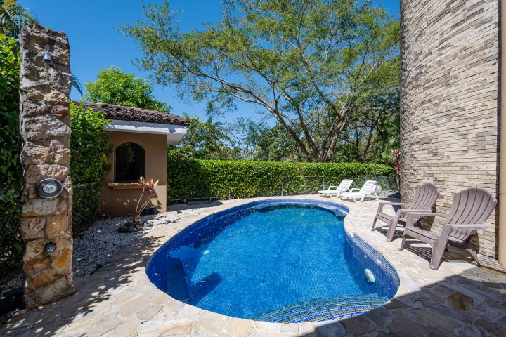 Small curved pool with lounge chairs, surrounded by stone patio and green foliage.