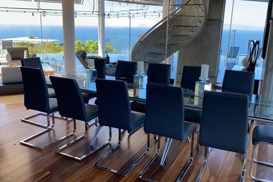 Modern dining room with glass table, dark chairs, spiral staircase, and ocean view.