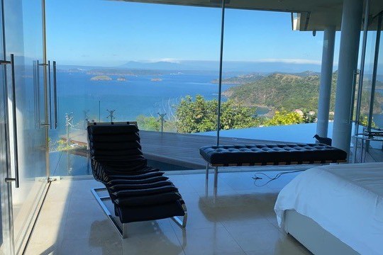 Modern room with glass walls, lounge chair, bed, and ocean view.