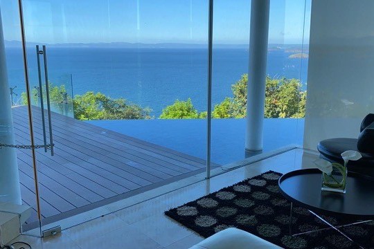 Modern room with ocean view, glass walls, infinity pool, and cozy seating area.