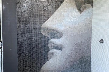 Shower with mosaic wall art of a classical statue face, dual showerheads, and a small bench.