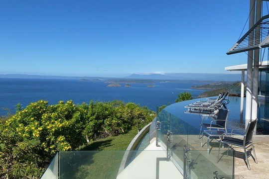 Coastal view from modern glass balcony with ocean, islands, and lush greenery under a clear blue sky.