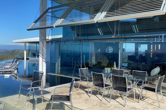 Modern patio with glass table, chairs, and pool in a luxurious setting overlooking a scenic landscape.