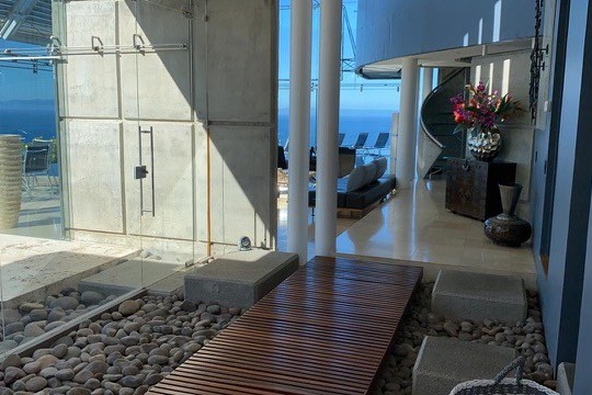 Modern interior with glass walls, stone path, wood walkway, and ocean view.