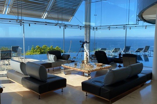 Modern living room with glass walls, ocean view, sofas, telescope, and a terrace with loungers.