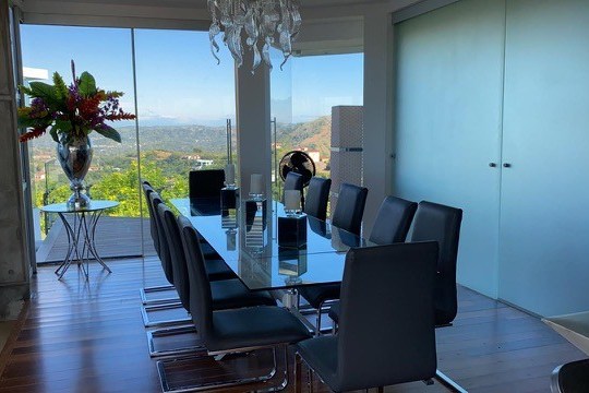 Modern dining room with glass table, black chairs, chandelier, and mountain view through large windows.
