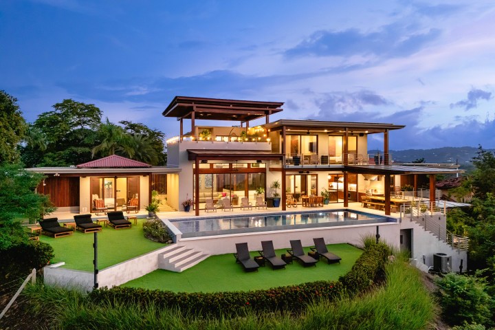 Modern hillside villa with pool at sunset, surrounded by lush greenery.