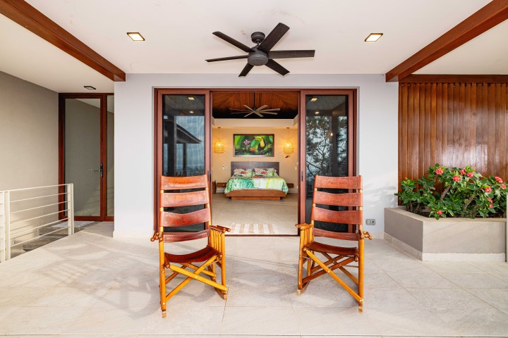 Two wooden chairs on a patio facing a bedroom with a ceiling fan and plants.