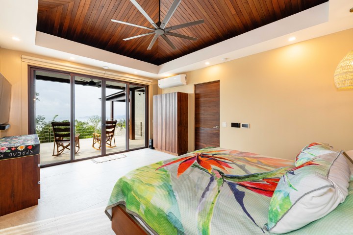 Bedroom with tropical-themed bedspread and sliding glass doors opening to a deck with rocking chairs.