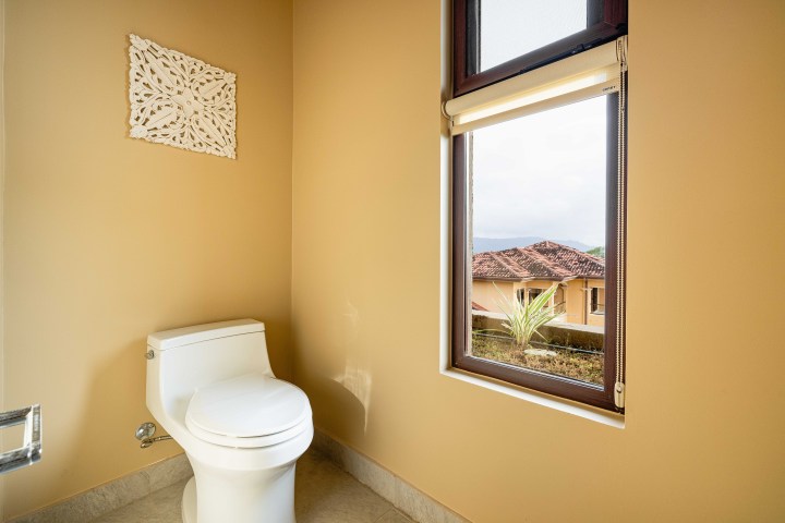 A toilet in a beige bathroom with a window showing a house with a red roof outside.