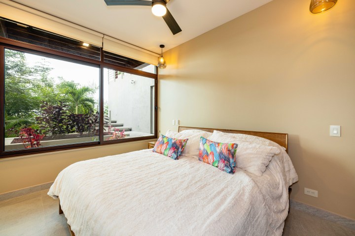 Modern bedroom with large window, ceiling fan, and a bed with colorful pillows.