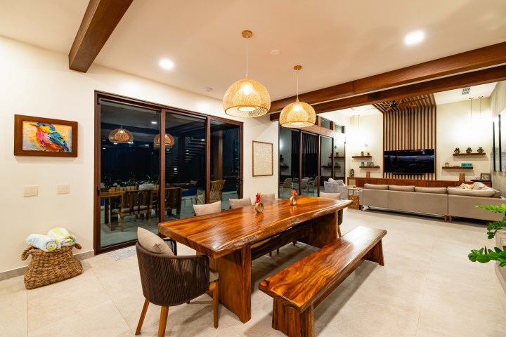Modern living area with wooden dining table, benches, pendant lights, and a large sofa in background.