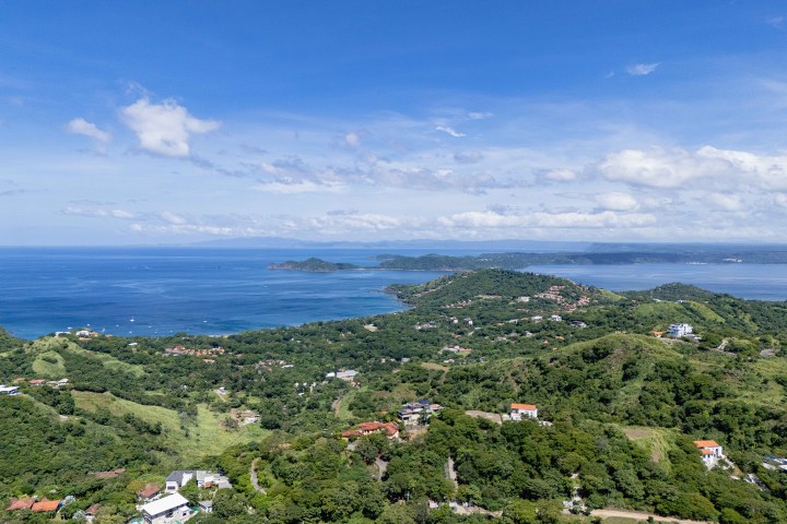 Aerial view of lush hills, scattered houses, and ocean with blue sky and clouds.