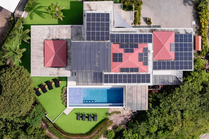 Aerial view of a modern house with solar panels and a pool, surrounded by greenery.