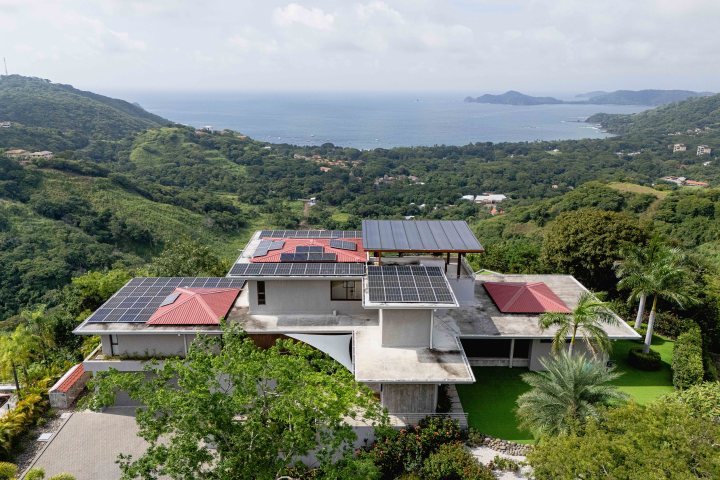 Modern house with solar panels on roof, green landscape, ocean view in background.